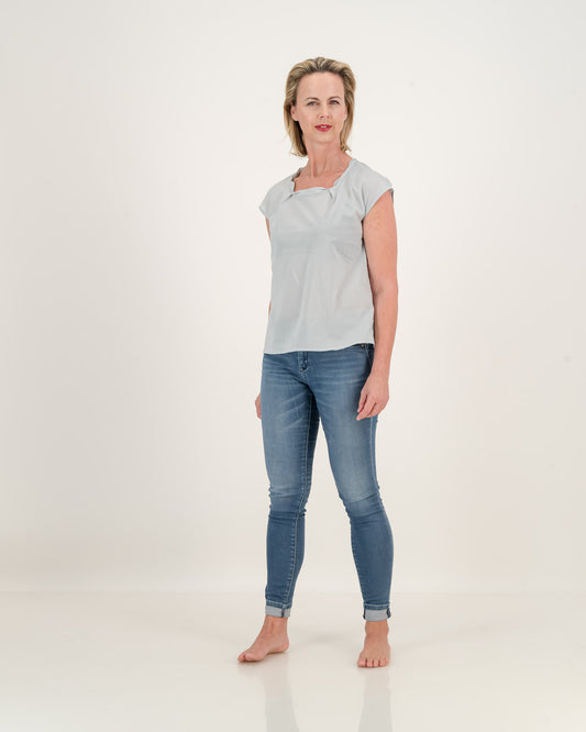 A woman wearing jeans and a subtle blue top. The top has a pleated neckline, adding elegance to its relaxed style. Stay cool and stylish in this lightweight cotton essential.