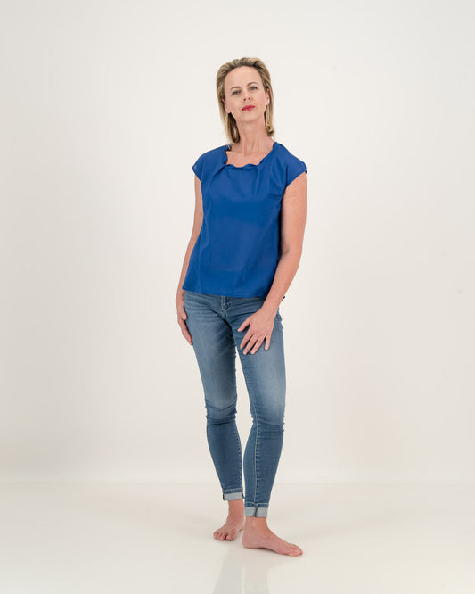 A woman wearing jeans and a relaxed sapphire blue top. The top has a pleated neckline, adding elegance to its relaxed style. Stay cool and stylish in this lightweight cotton essential.