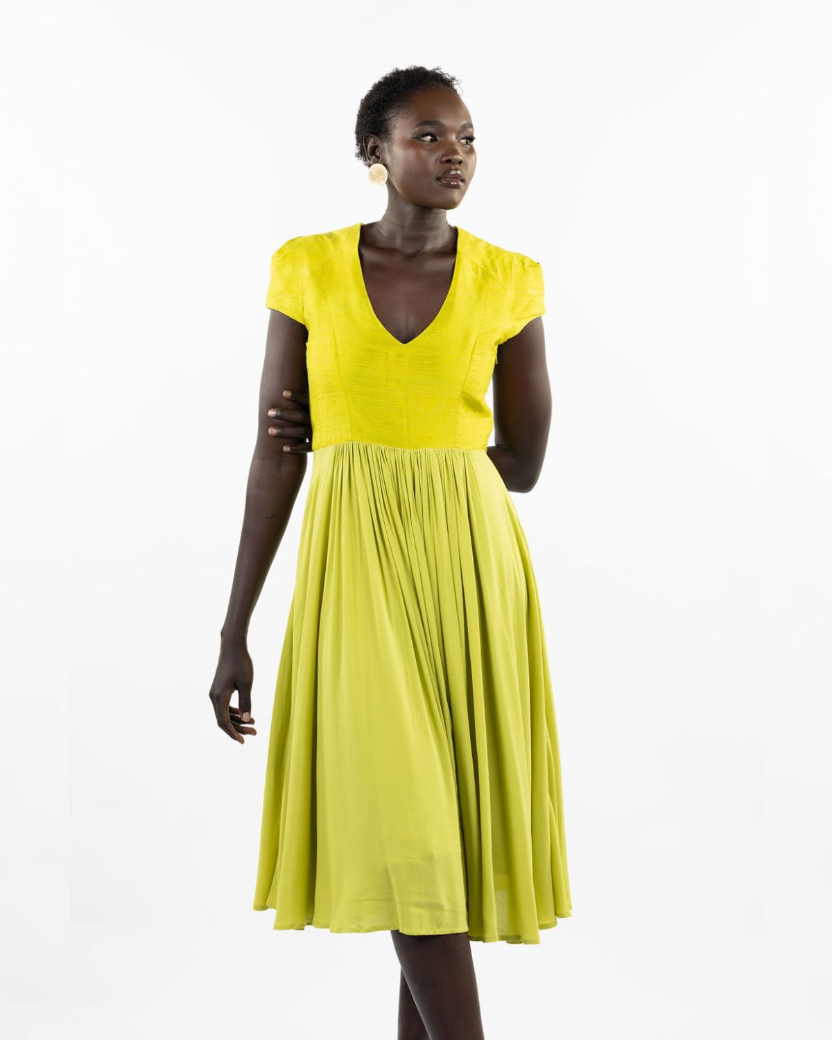 A woman models a chartreuse-colored knee-length dress with a gathered skirt and a V-neckline. The dress features cap sleeves and a flattering waist cinch.