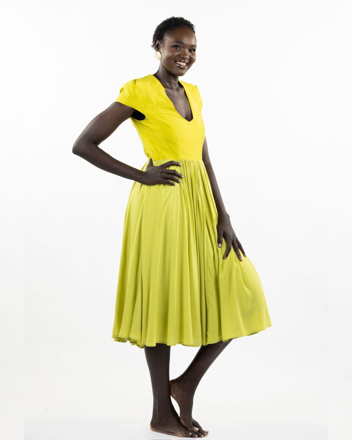 A woman models a chartreuse-colored knee-length dress with a gathered skirt and a V-neckline. The dress features cap sleeves and a flattering waist cinch.