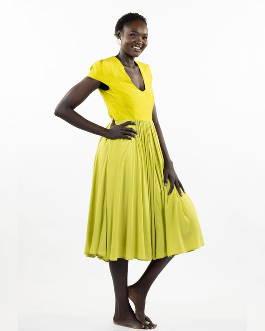 A woman models a chartreuse-colored knee-length dress with a gathered skirt and a V-neckline. The dress features cap sleeves and a flattering waist cinch.