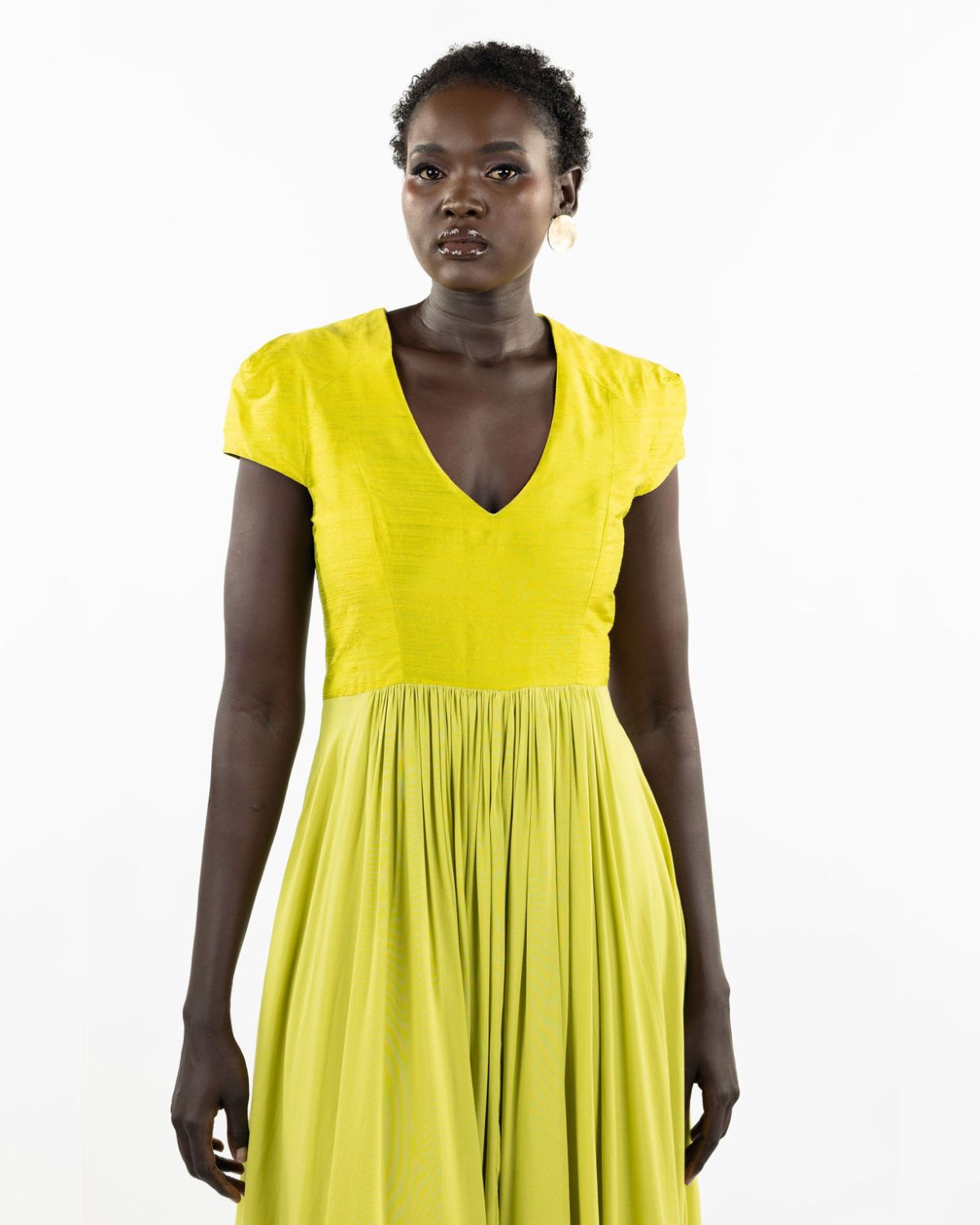A woman models a chartreuse-colored knee-length dress with a gathered skirt and a V-neckline. The dress features cap sleeves and a flattering waist cinch.