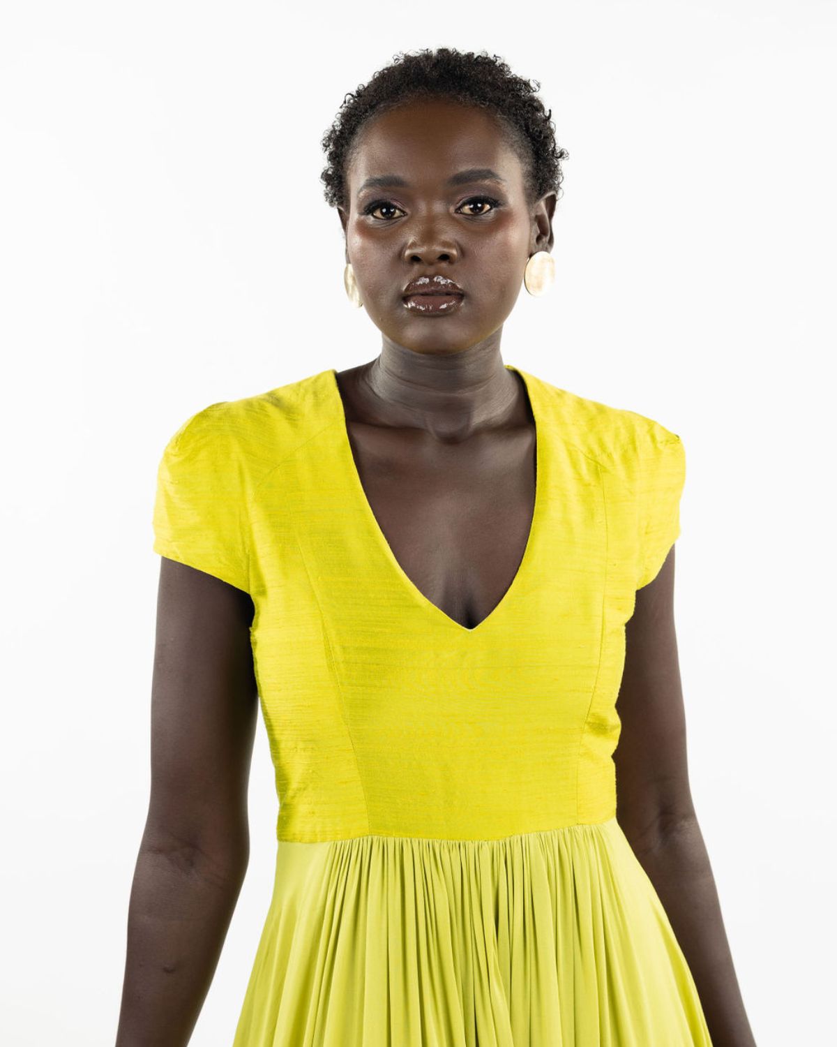 A woman models a chartreuse-colored knee-length dress with a gathered skirt and a V-neckline. The dress features cap sleeves and a flattering waist cinch.