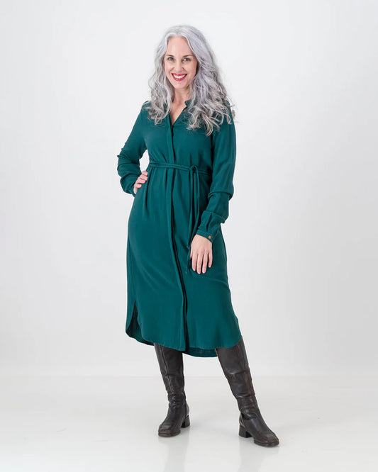 A stunning older woman with long silver hair wears the emerald green imogen dress from LUNAR. The dress is below the knee and has a cinched in waist with a long sleeve and hidden button-stand.