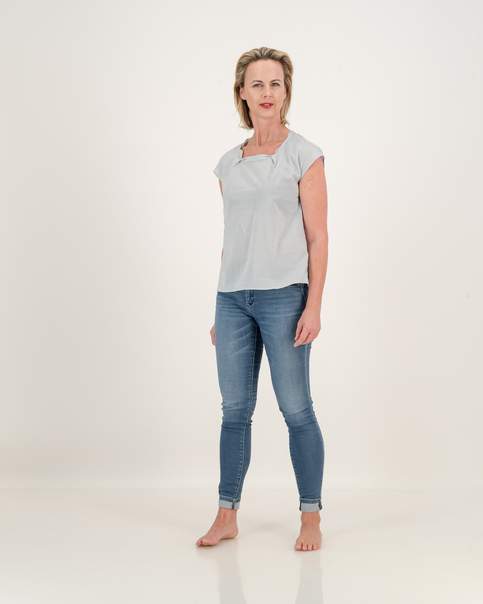 A woman wearing jeans and a subtle blue top. The top has a pleated neckline, adding elegance to its relaxed style. Stay cool and stylish in this lightweight cotton essential.
