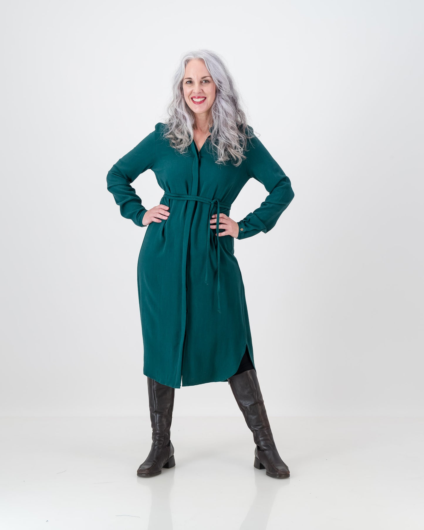 A stunning older woman with long silver hair wears the emerald green imogen dress from LUNAR. The dress is below the knee and has a cinched in waist with a long sleeve and hidden button-stand.