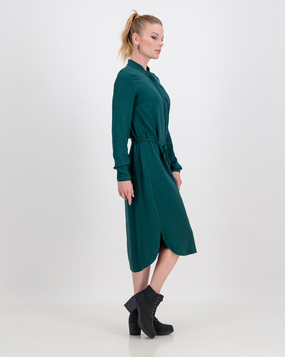 A young vibrant woman wears the imogen dress is and is facing the side so that we can see the shape of the emerald green imogen dress. It has a below the knee hem line and a concealed button stand. The dress also features long sleeves that can be folded or ruched up.