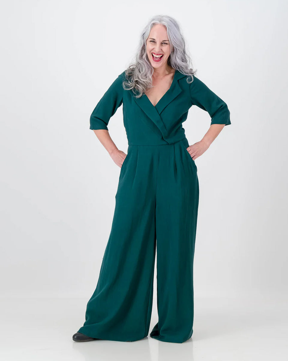 A woman standing with hands on hips, wearing an emerald green jumpsuit with an asymmetrical collar, wide-leg pants, and pockets.