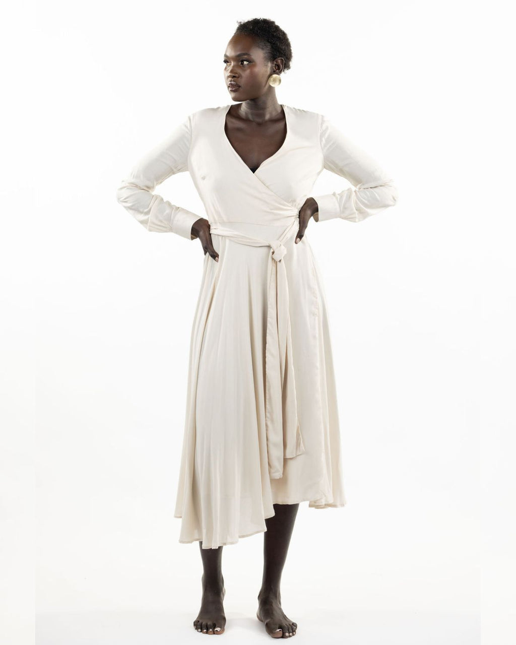 A model wearing a stone-colored eva wrap dress with a v-neckline, long sleeves, covered buttons, and an asymmetrical hem.