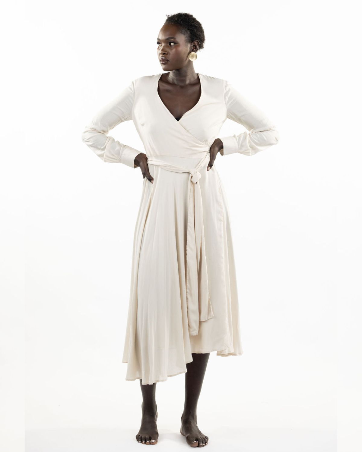 A model wearing a stone-colored eva wrap dress with a v-neckline, long sleeves, covered buttons, and an asymmetrical hem.