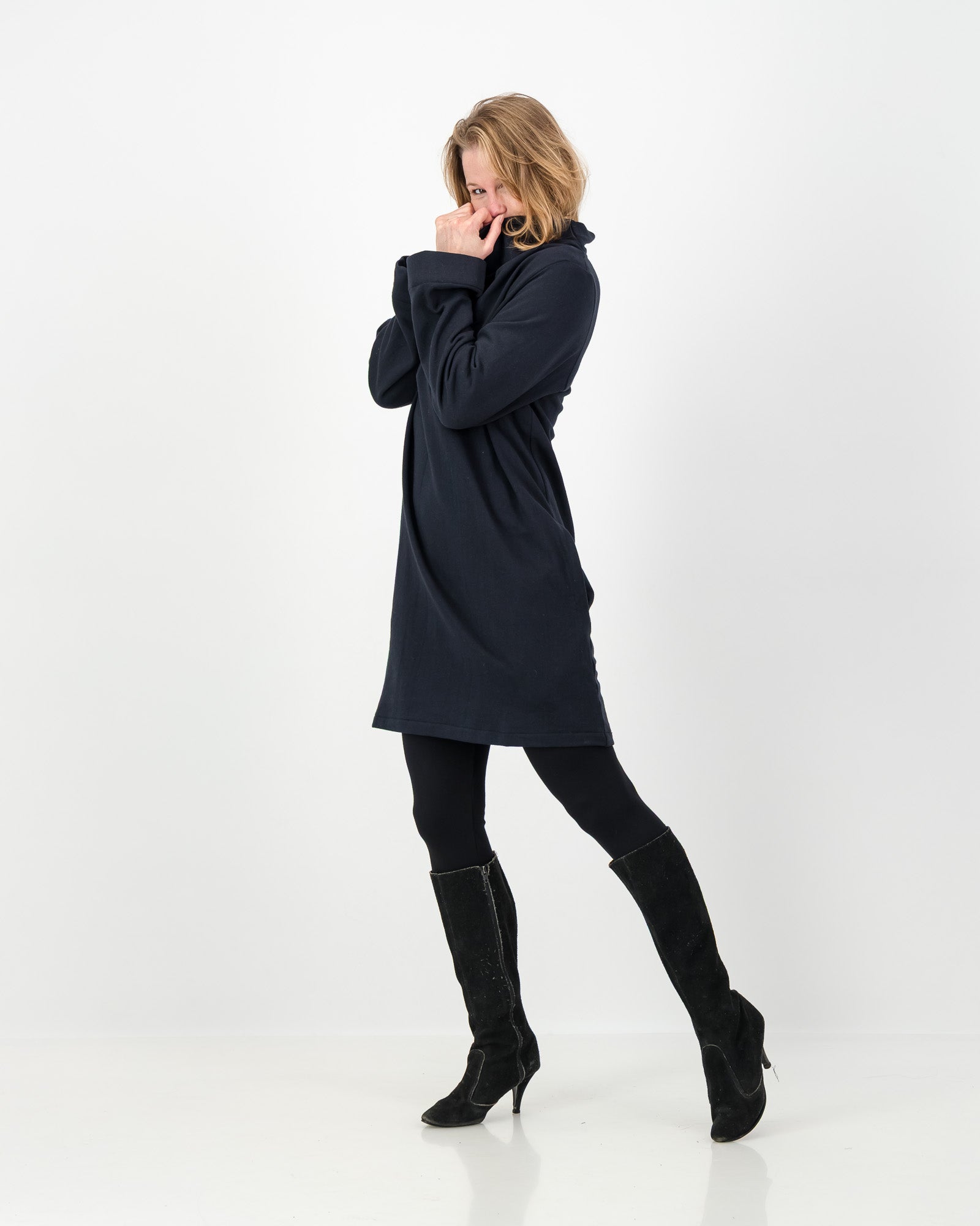 Sideview of a black-colored winter dress with a cowl neckline, full-length sleeves, and a straight cut, worn by a lady with black boots and leggings.