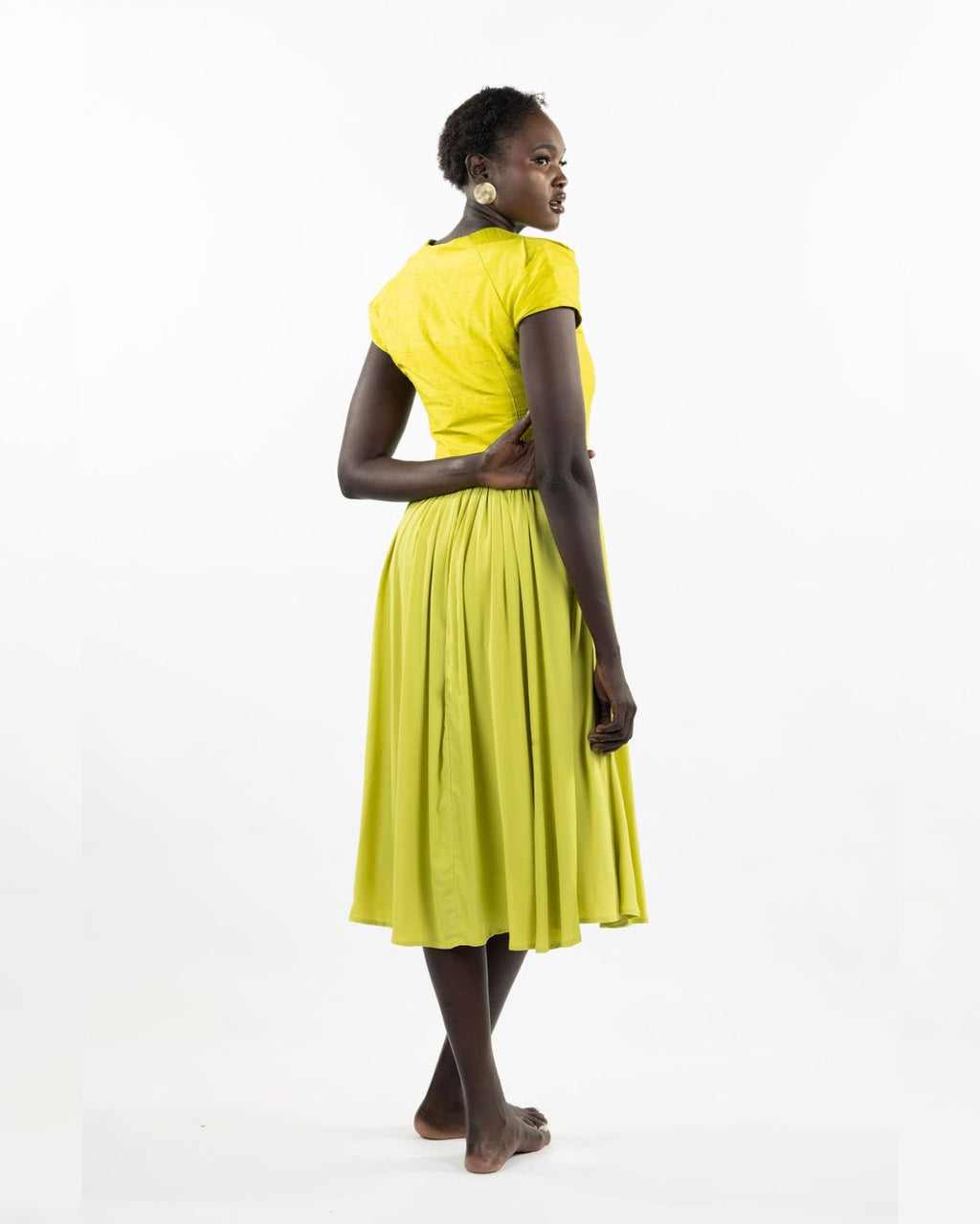 A woman models a chartreuse-colored knee-length dress with a gathered skirt and a V-neckline. The dress features cap sleeves and a flattering waist cinch.