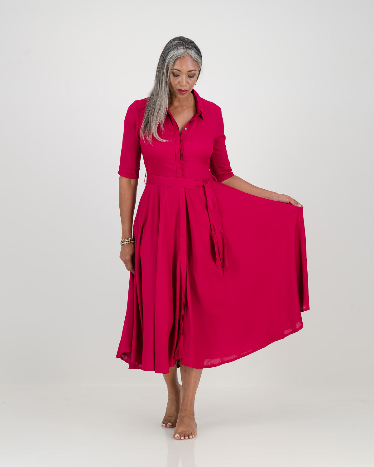 Woman wearing chic cerise Abigail dress with 3/4 sleeves, collar, and belt at waist. Features full skirt, concealed buttons, and pockets. Elegant and versatile.